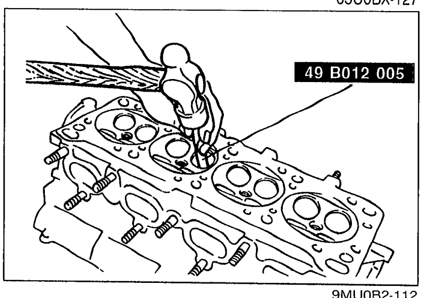 Removing the valve guide from the cylinder head (top, combustion-chamber side facing down) using SST 49 B012 005 driven through the guide from the spring-seat side.