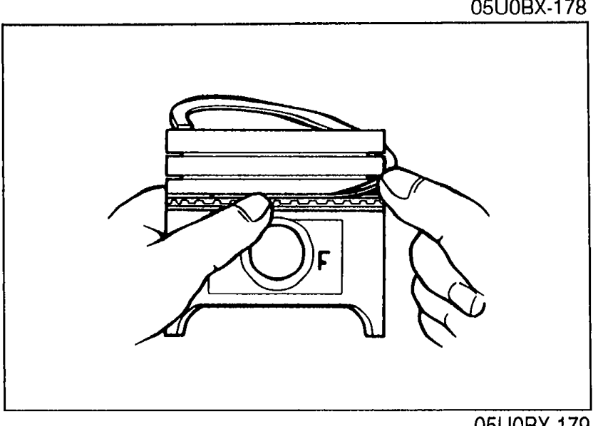 Hand installing the three-piece oil ring (oil ring spacer and two rails) onto a piston. The piston has an 'F' mark visible indicating Front orientation.