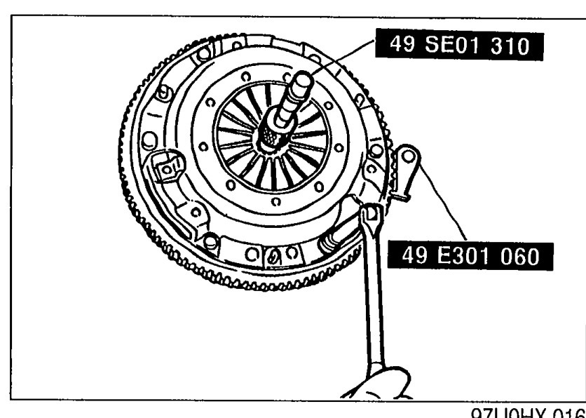 Photo of the clutch cover installed on the flywheel with SST 49 SE01 310 (clutch disc centering tool) inserted through the disc, and SST 49 E301 060 (ring-gear brake) engaged on the ring gear teeth to hold the assembly while loosening cover bolts.