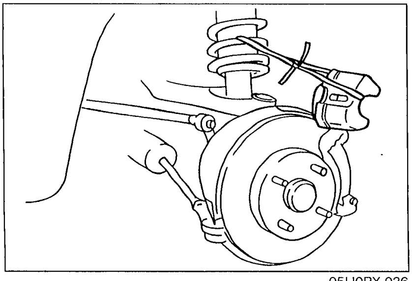 Photograph of the front strut/knuckle/brake-caliper area showing the caliper rotated upward off the rotor (with lower lock bolt removed) and the rotor exposed for pad removal.